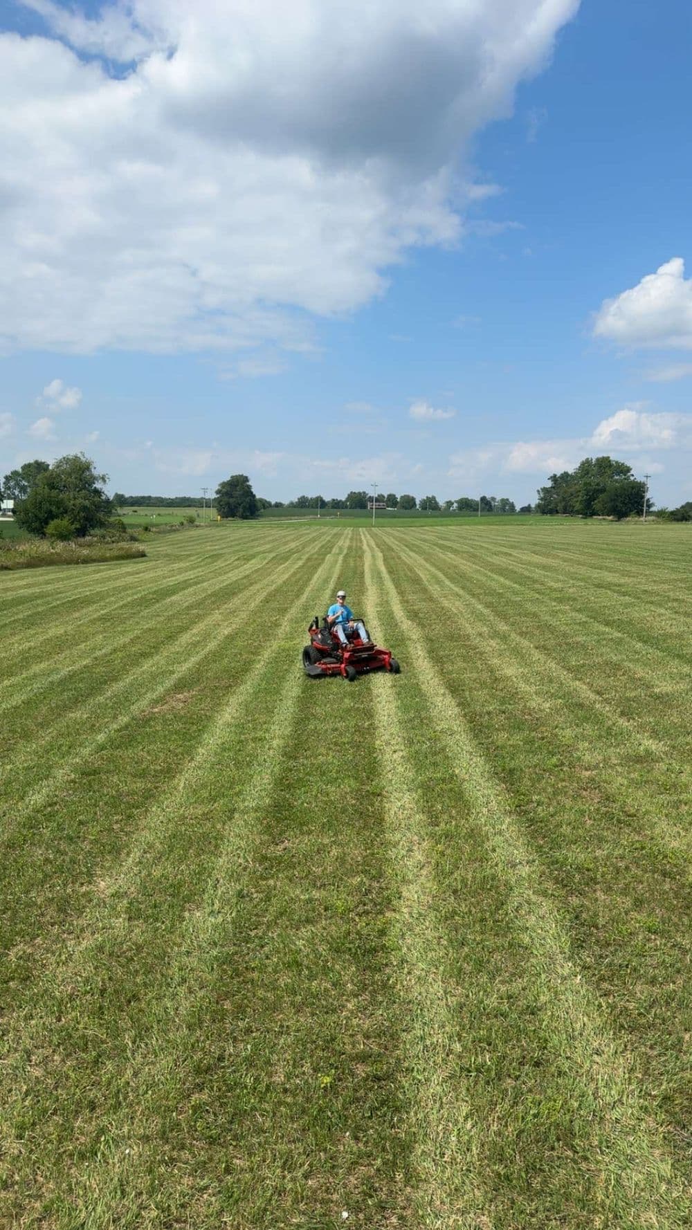 Person mowing a grassy field with a riding lawn mower under a blue sky and scattered clouds.