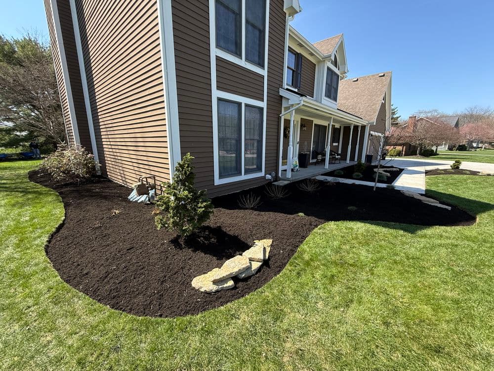 Mulched landscape around a brown house featuring grass, shrubs, and a stone pathway.