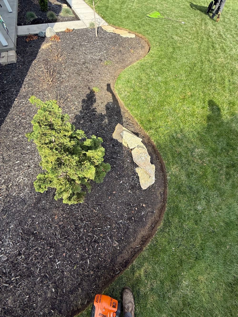 Landscaped garden with a curved mulched border, green grass, and a small shrub by a large rock.