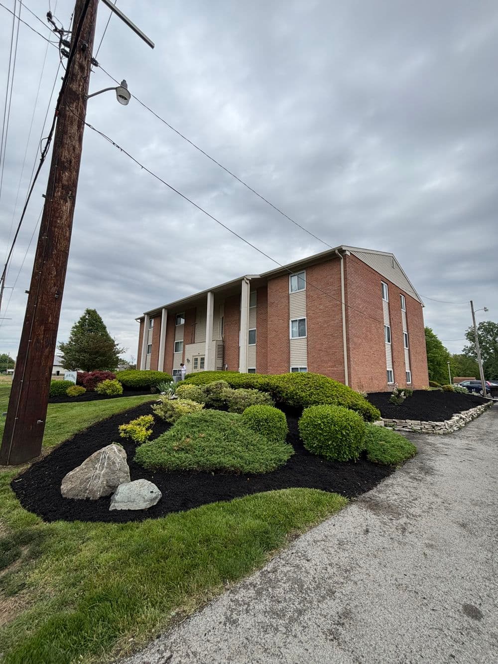 Brick apartment building with landscaped gardens and black mulch under a cloudy sky.