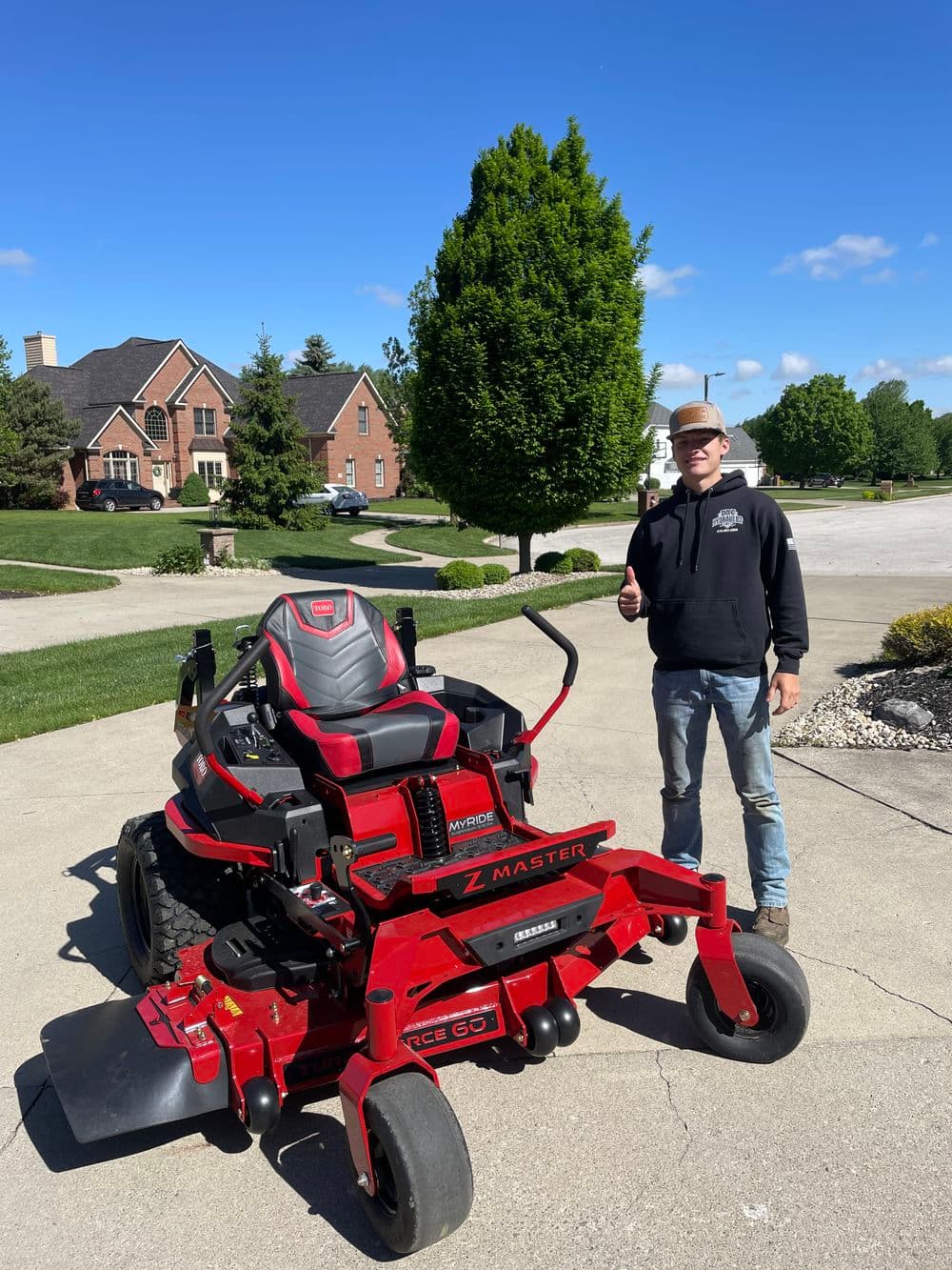 Man giving thumbs up next to a red zero-turn mower on a sunny residential street.