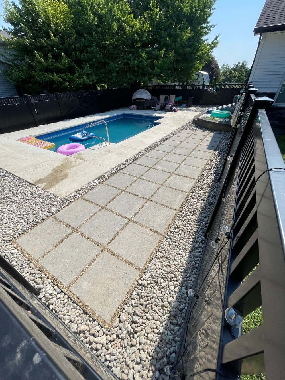 Modern backyard pool area with paved walkway, lounge chairs, and green trees.