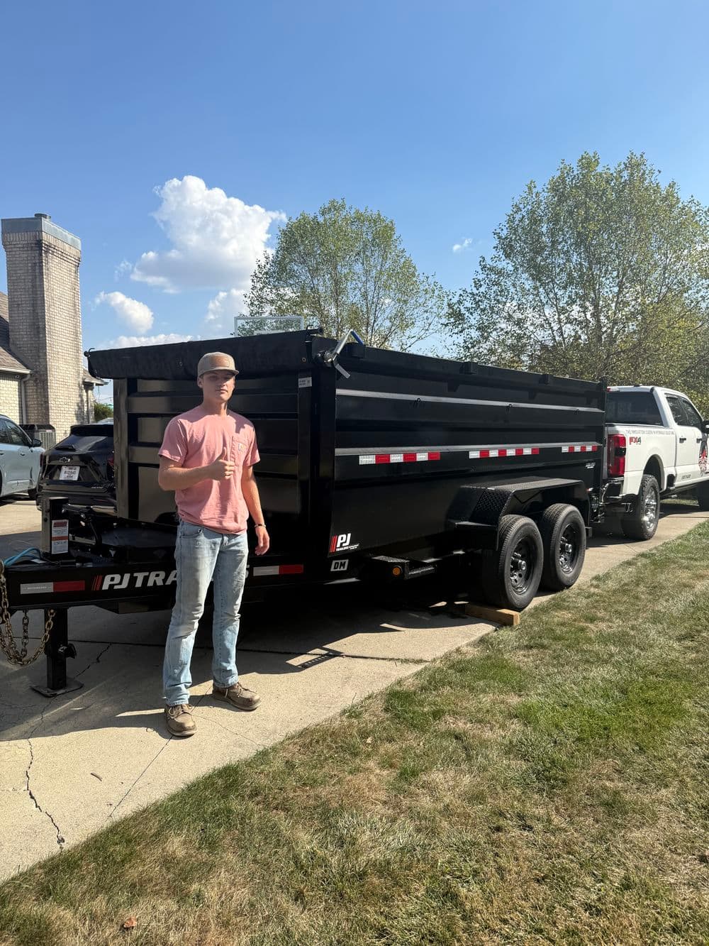 Person standing next to a PJ Trailers dump trailer parked in a driveway.