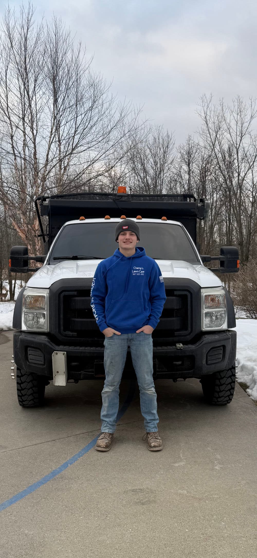 Young man in a blue hoodie stands in front of a pickup truck on a snowy driveway.
