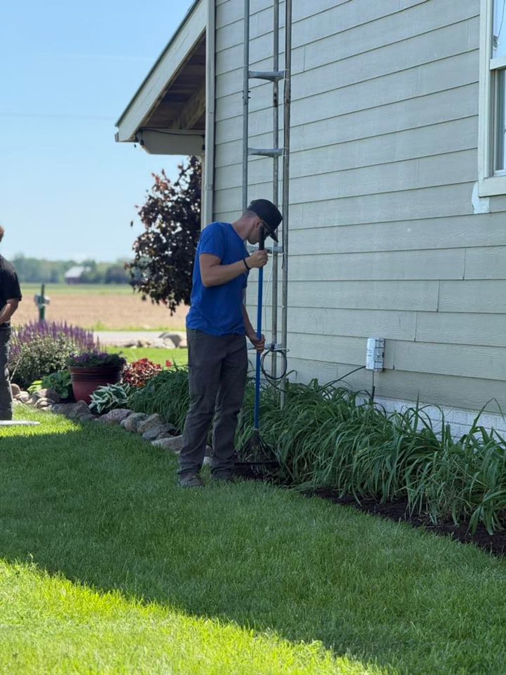 Man using a tool to tend to garden landscaping by a house on a sunny day.