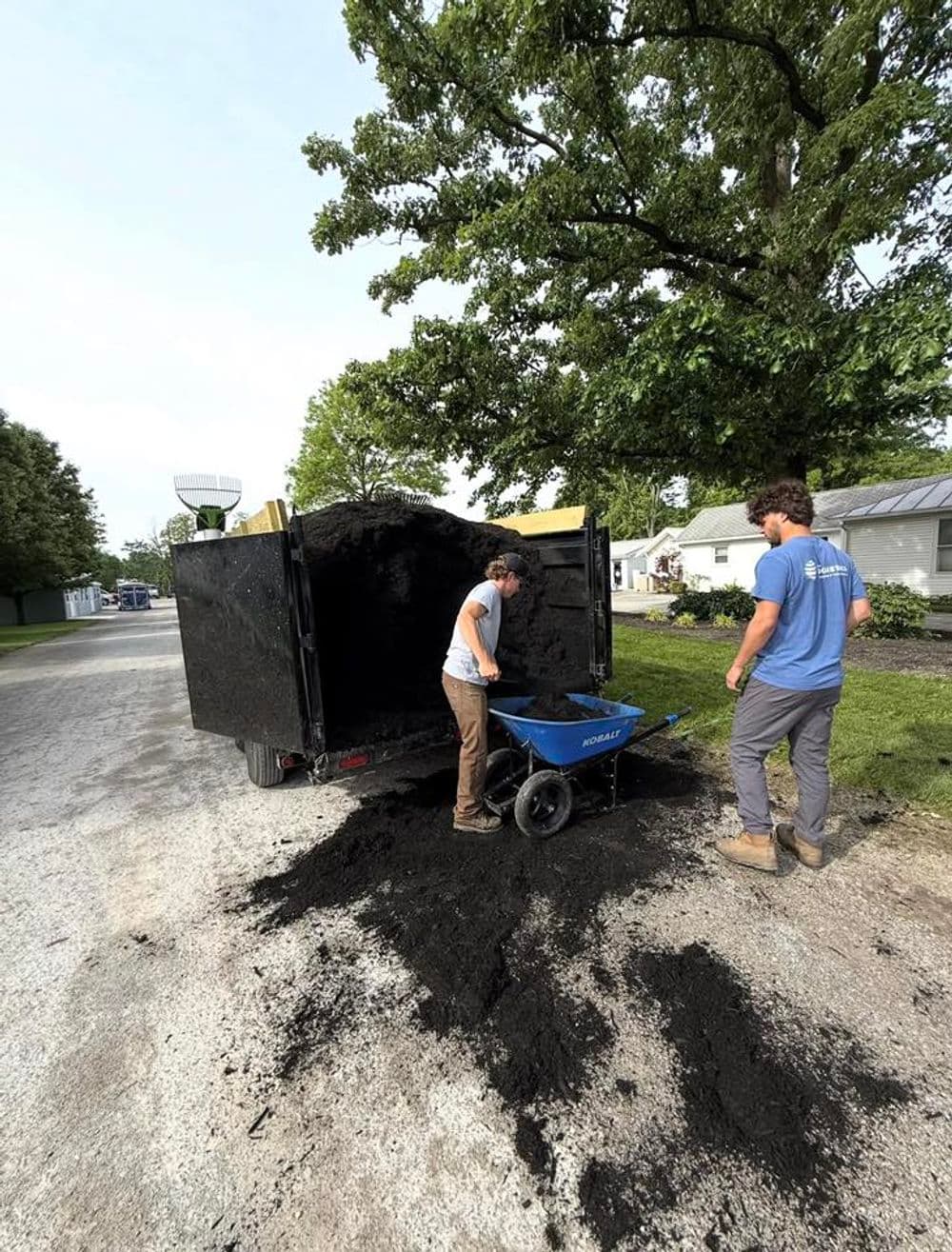 Workers unloading soil from a trailer into a wheelbarrow on a gravel road.